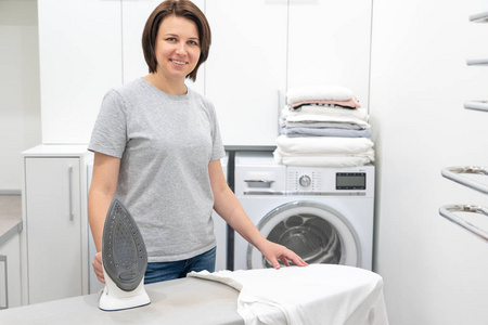 一位女士微笑着站在洗衣房的熨衣板旁，背景是洗衣机