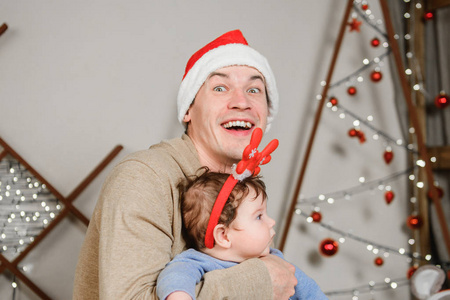 爸爸和儿子在圣诞树前拥抱。一个快乐的孩子和他的爸爸正在看镜框。爸爸和儿子拥抱过圣诞节。玩得开心。头上有角。新年帽。