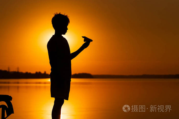 橙色的夕阳下 一个十几岁的男孩看着手中的纸飞机 一个小孩拿着折纸飞机在水面上的摇篮 照片 正版商用图片26jdtm 摄图新视界