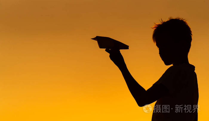 一个男孩站在橙色的夏日日落背景下的黑色剪影手里拿着小纸飞机水平