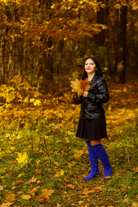 一个年轻的黑发女人穿着蓝色靴子，手里拿着黄叶走在秋天的公园里