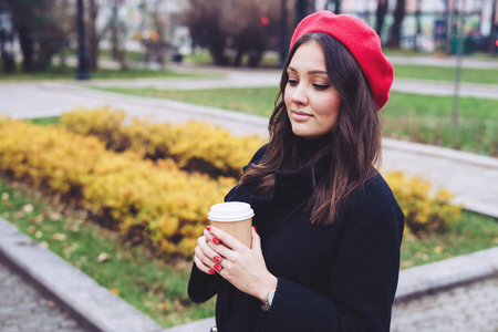 在秋天的公园里，女人拿着黑色外套和红色贝雷帽的饮料。