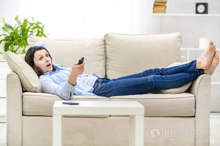 震惊和惊讶的女人张开嘴一边看电视躺在沙发上复制空间
