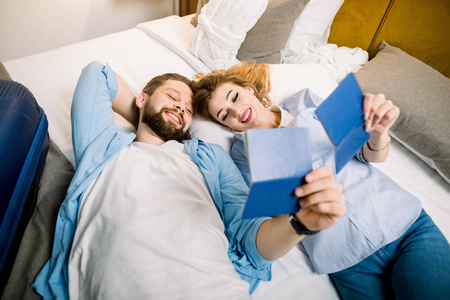 微笑的年轻夫妇躺在酒店或公寓的床上，看着他们的旅行护照