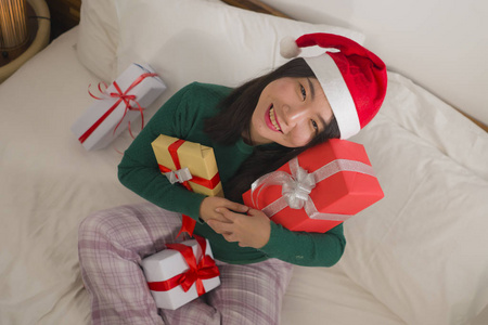 圣诞生活方式画像年轻美丽快乐的亚裔日本妇女在床上戴着圣诞老人的帽子，手里拿着圣诞礼物和礼品盒，兴奋而欢快