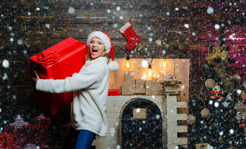 雪地里的女人。魅力庆祝新年。金发的圣诞老人妇女拿着礼品盒在复古墙上。穿着圣诞老人衣服的漂亮女孩的肖像。戴着红色圣诞帽的女人拿着礼