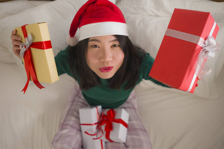 圣诞生活写真年轻美丽快乐的亚裔中国妇女在床上戴着圣诞老人的帽子，手里拿着很多圣诞礼物和礼品盒，兴奋而欢快