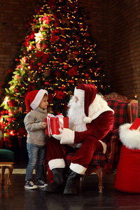 圣诞老人和带礼物的小男孩在室内的圣诞树旁