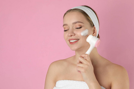 年轻女子用刷子和洁面泡沫在粉色b上洗脸