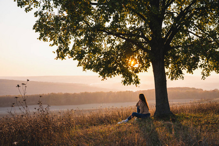 风景树下的美女穿着休闲服的漂亮女孩坐在大树下看书,看着美丽的日落
