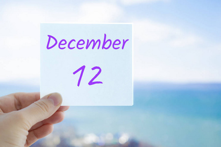 12月12日。手持贴纸与文字12月12日在模糊的背景上的海洋和天空。复制文本空间。月历概念