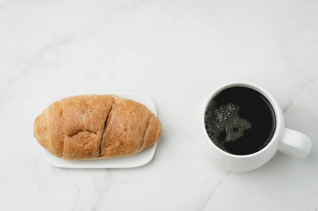 White mug with black coffee and croissant on white stone table. 