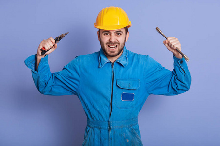 这是一个愤怒的建筑工人工程师的特写镜头，他戴着黄色的头盔和蓝色的制服，双手举起手来，愤怒地喊着什么，大胡子的家伙双手拿着扳手工具