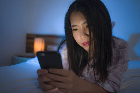 年轻美丽快乐甜美的亚洲韩国女人,穿着睡衣,晚上在床上享受手机应用