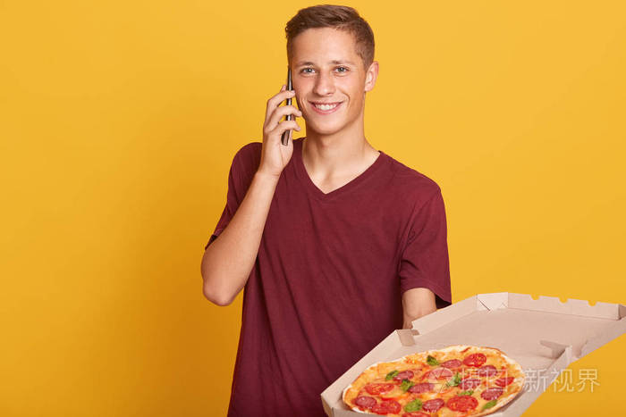 一位快乐的男性快递员通过他的智能手机查看到客户家的路线，拿着打开的装有比萨饼的纸箱，穿着休闲的勃艮第t恤，在黄色的工作室墙上摆出