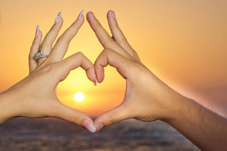 浪漫的情侣在夕阳下用手指指点着一颗心.手形成一个心形与日落剪影.
