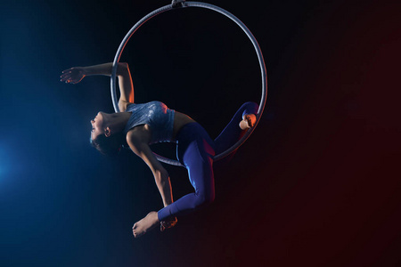 Young woman performing acrobatic element on aerial ring against 