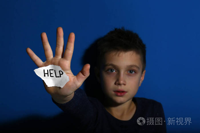 Abused little boy with sign HELP near blue wall, focus on hand. 