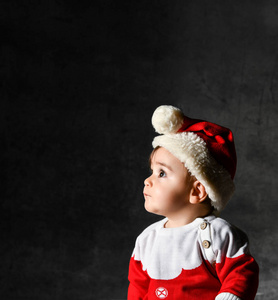 一个穿着红色毛衣，戴着圣诞帽的小男孩，坐在黑暗的背景上。