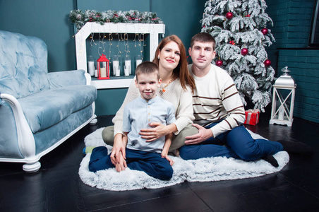 一家人，妈妈，爸爸和儿子坐在圣诞树旁的地板和壁炉上。