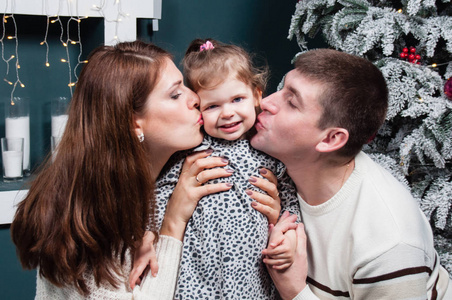 一对恩爱夫妻，一对英俊的男人和一对漂亮的女人在圣诞树旁亲吻他们的小女儿的脸颊