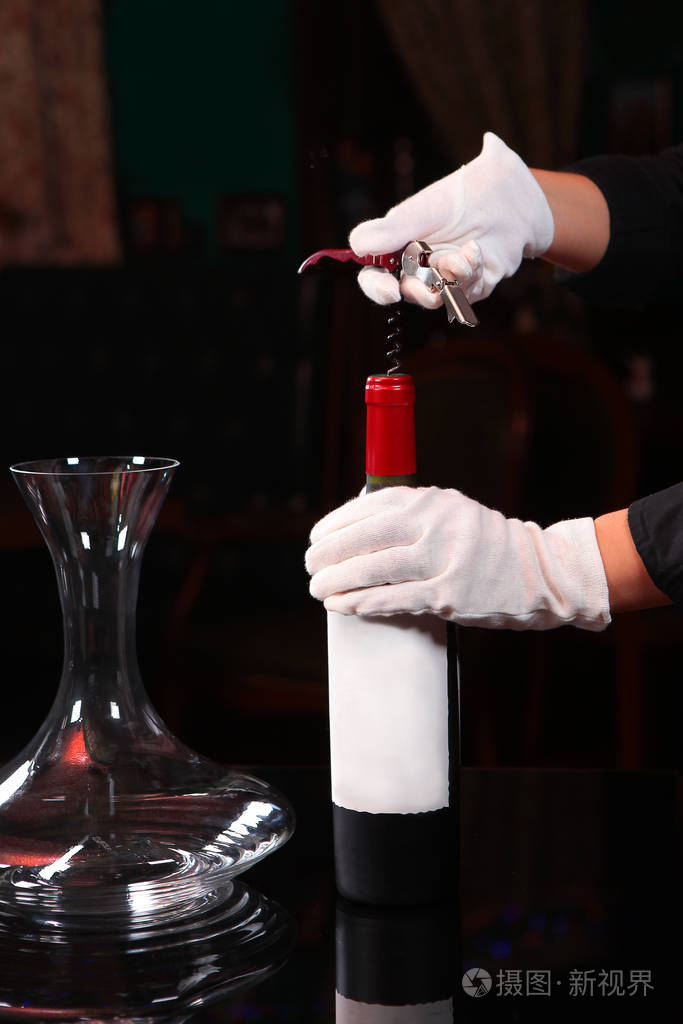 侍者戴着白手套的手用开瓶器打开一瓶红酒在餐厅内部的一张深色背景