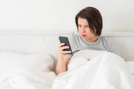 惊讶的女人坐在铺着羽绒被的床上用智能手机