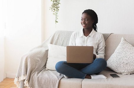 非洲女人坐在沙发上，带着笔记本电脑，在家里上网工作