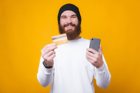 一个快乐的大胡子时髦在黄色背景上，手里拿着信用卡和智能手机