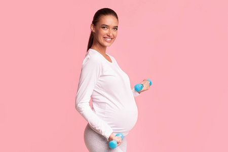 快乐的怀孕女孩举着哑铃站在粉红色的工作室背景上