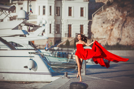 穿着飘逸的红色长裙的模特儿开着游艇沿着海滨散步