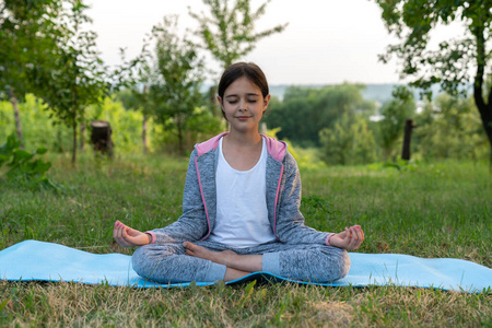 户外前景色全身肖像一个放松的黑发女孩穿着灰色运动服练习瑜伽坐在花园中的瑜伽垫上