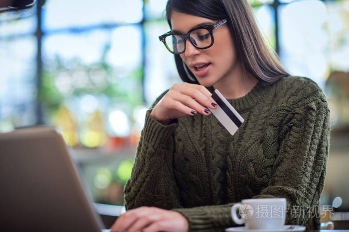 穿着毛衣的白种黑发美女坐在咖啡馆里用键盘打字手里拿着信用卡