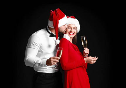 这对幸福的夫妇戴着圣诞老人的帽子，在黑暗的背景下喝着香槟