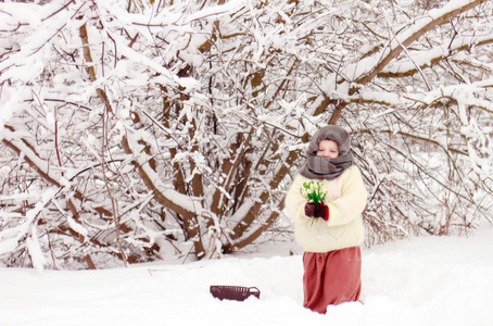 一个悲伤的女孩带着雪滴在雪林里,穿着温暖的白色披肩,棕色裙子和篮子