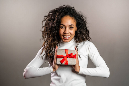 年轻的混血女孩微笑着拿着礼品盒，背景是灰色的工作室。美国黑人女性的可爱肖像。圣诞气氛