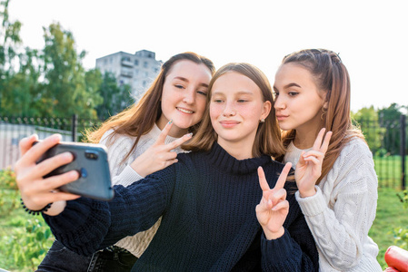 3女朋友1415岁青少年，自拍手机，秋季公园，大学放学后放松，观看视频社交网络智能手机屏幕。休闲装，毛衣牛仔裤。