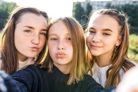 3名女生女友青少年，1415岁，在电话相机上自拍，秋日在城市街头，大学和学校下课休息。穿日常服装毛衣。