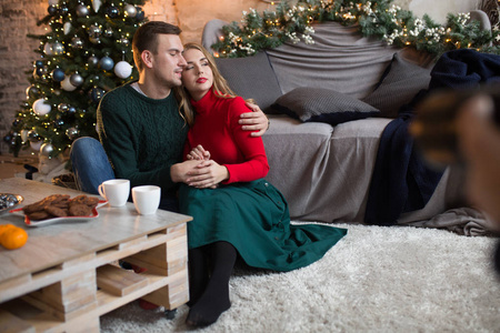 一对幸福相爱的夫妇坐在沙发旁边，旁边是一棵圣诞树，一张桌子和橘子，杯子里放着巧克力，咖啡加牛奶，可可，拥抱。家里的温柔和舒适。