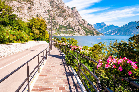 舒适的城市街道Limone Sul Garda，铺路石人行道，金属栏杆上盛开的花朵，背景是令人惊叹的加尔达湖和白云石山脉的树木。