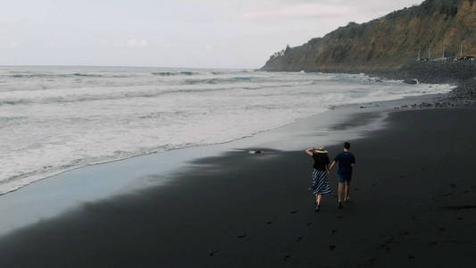 漂亮的电影镜头。一对年轻夫妇挽手沿着黑色火山沙滩奔跑。西班牙特内里费鸟瞰图