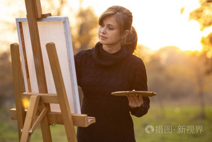 年轻漂亮的女画家在画架上画画女孩用画笔和调色板在秋天的风景背景上