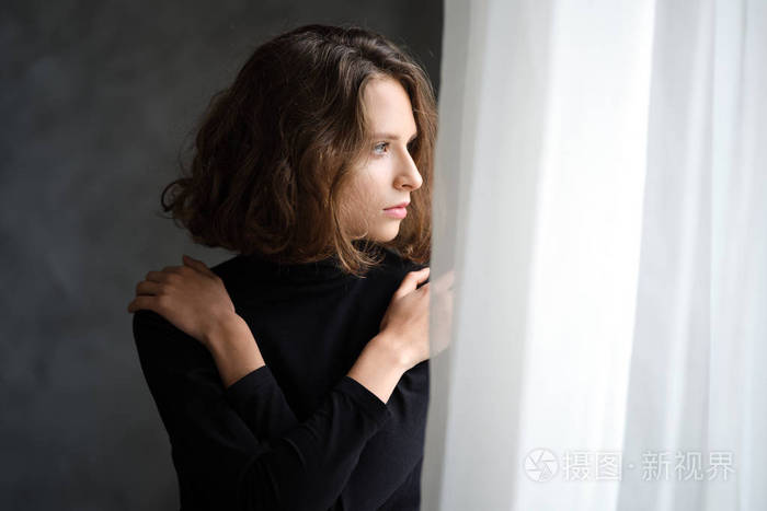 美丽的女孩抱住自己的肩膀若有所思地看着窗外