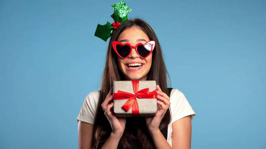 美丽的年轻女孩微笑着拿着礼品盒，背景是蓝色的工作室。戴着圣诞帽的可爱肖像。圣诞气氛。