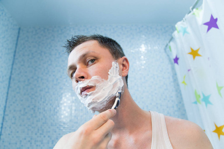 一个男人用剃须刀在泡沫中刮脸.男人在镜子前刮胡子照片