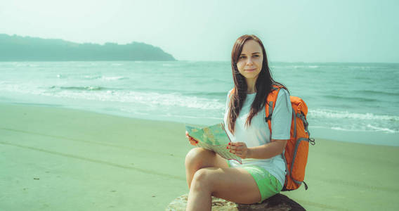 女游客在海边看地图。赤脚女人背着背包坐在浮木上，在波涛汹涌的大海附近的沙滩上休息