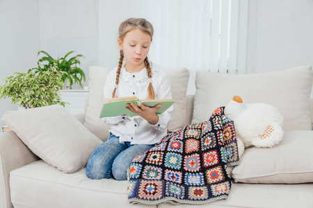 可爱的小女孩蜷着腿坐着,膝盖上放着书,毛毯下有泰迪熊.