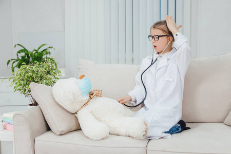 穿着医生外套的漂亮小女孩，蜷着腿坐在沙发上，用听诊器检查她生病的泰迪熊。
