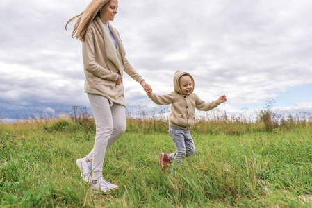 年轻的女人妈妈牵着小男孩在街头公园散步，穿着暖和的衣帽，草云背景。玩得开心开心快乐。周末漫步大自然。情感幸福。
