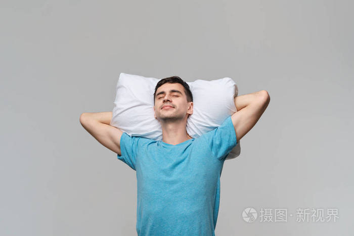 穿着蓝色t恤的疲惫的黑发男人躺在一个白色枕头上枕头被隔离在灰色的